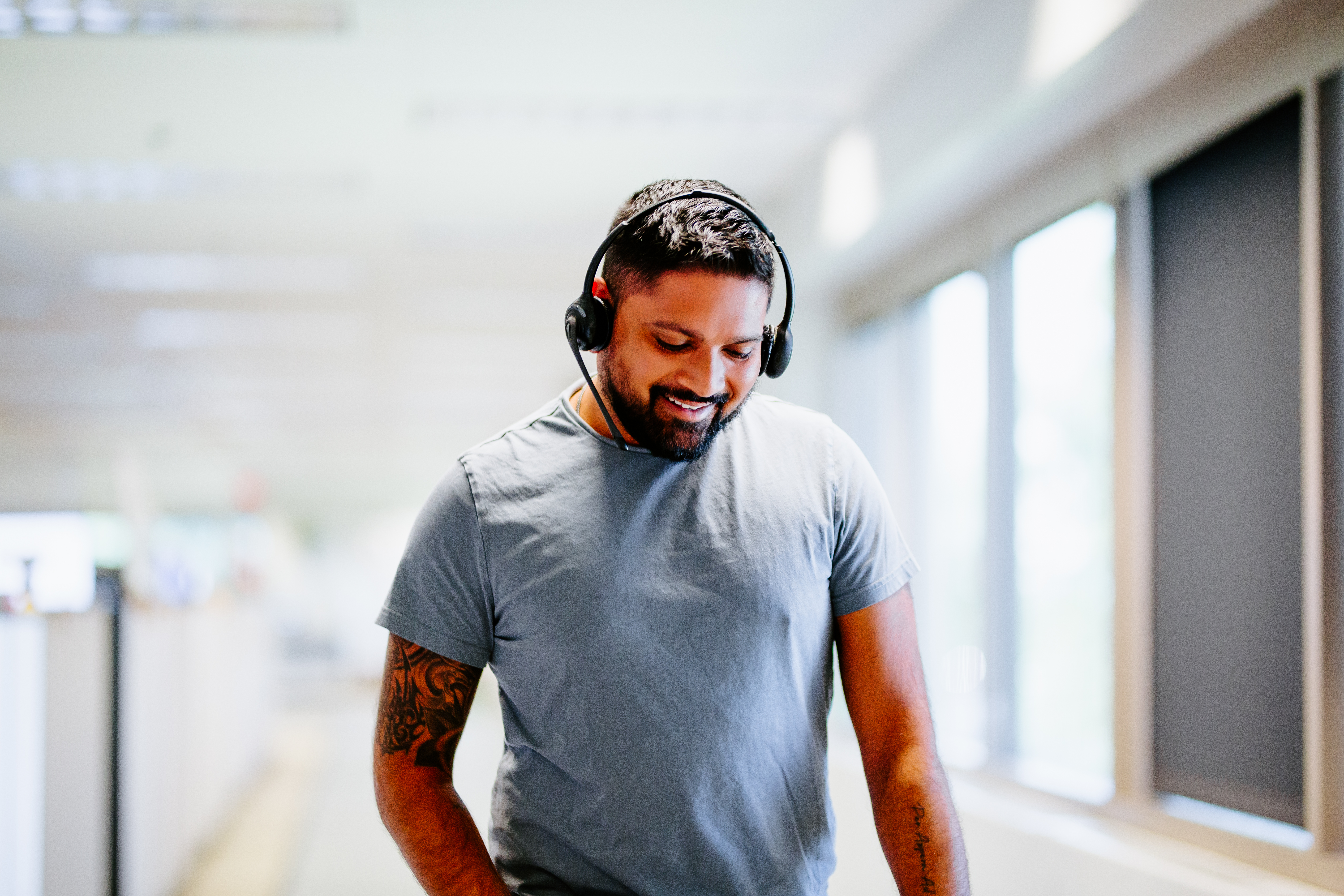 Man with dark hair, beard and tattoos on his right arm talking on a phone headset while walking down the hall