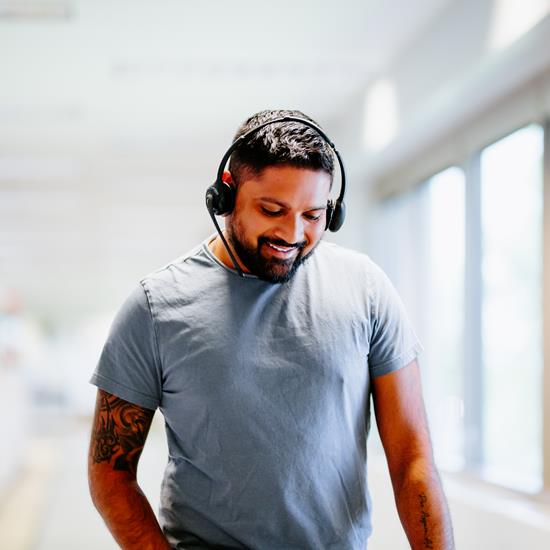 Man with dark hair, beard and tattoos on his right arm talking on a phone headset while walking down the hall