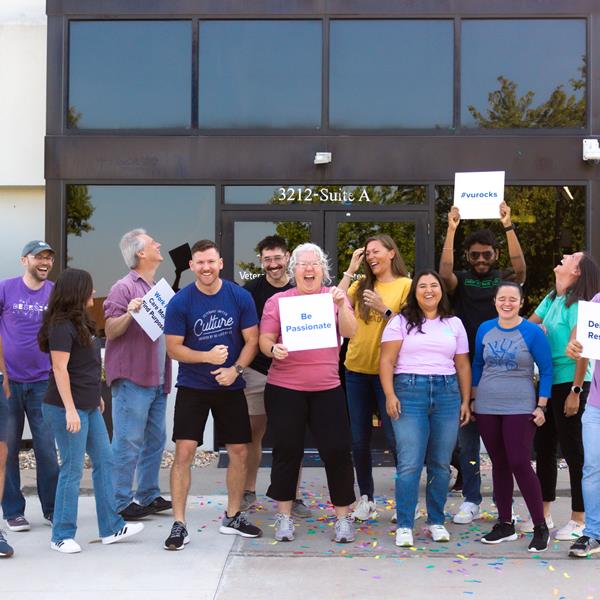 Group of employees outside smiling, cheering and holding signs with VU Values written on them