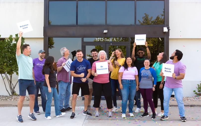 Group of employees outside smiling, cheering and holding signs with VU Values written on them